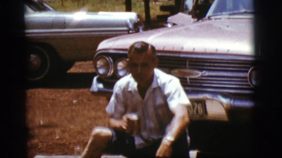 The man turns more toward the camera while seated with a can, with classic parked cars filling the background.
