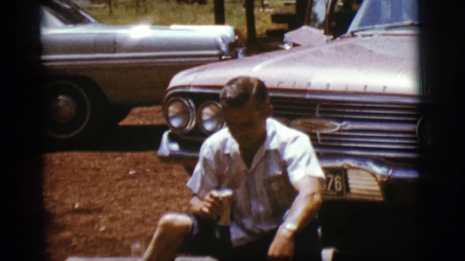 A seated man looks down while holding a can beside a 1960s car with chrome grille and dual headlights.