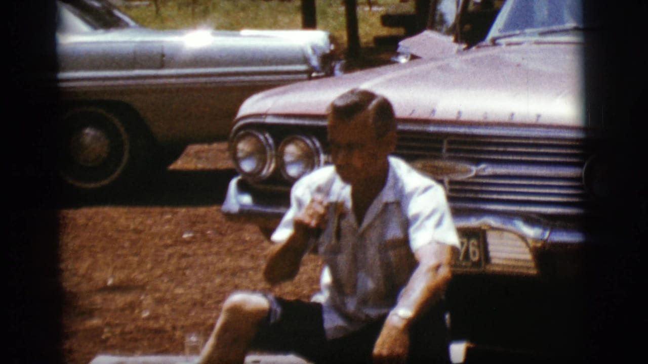 Man seated on the ground holding a can in front of a vintage car.