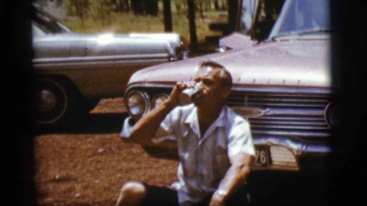 Man seated in front of a vintage car drinking from a can.
