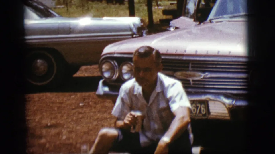 The man sits in front of the car with the drink can lowered, framed by another parked car and trees in the background.