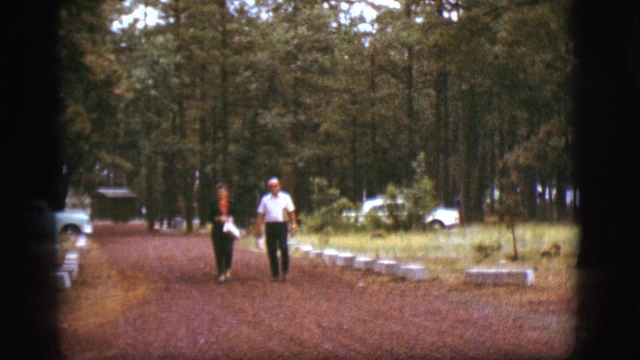 Man and woman walk toward the camera on a reddish dirt path lined with trees and low curb markers.