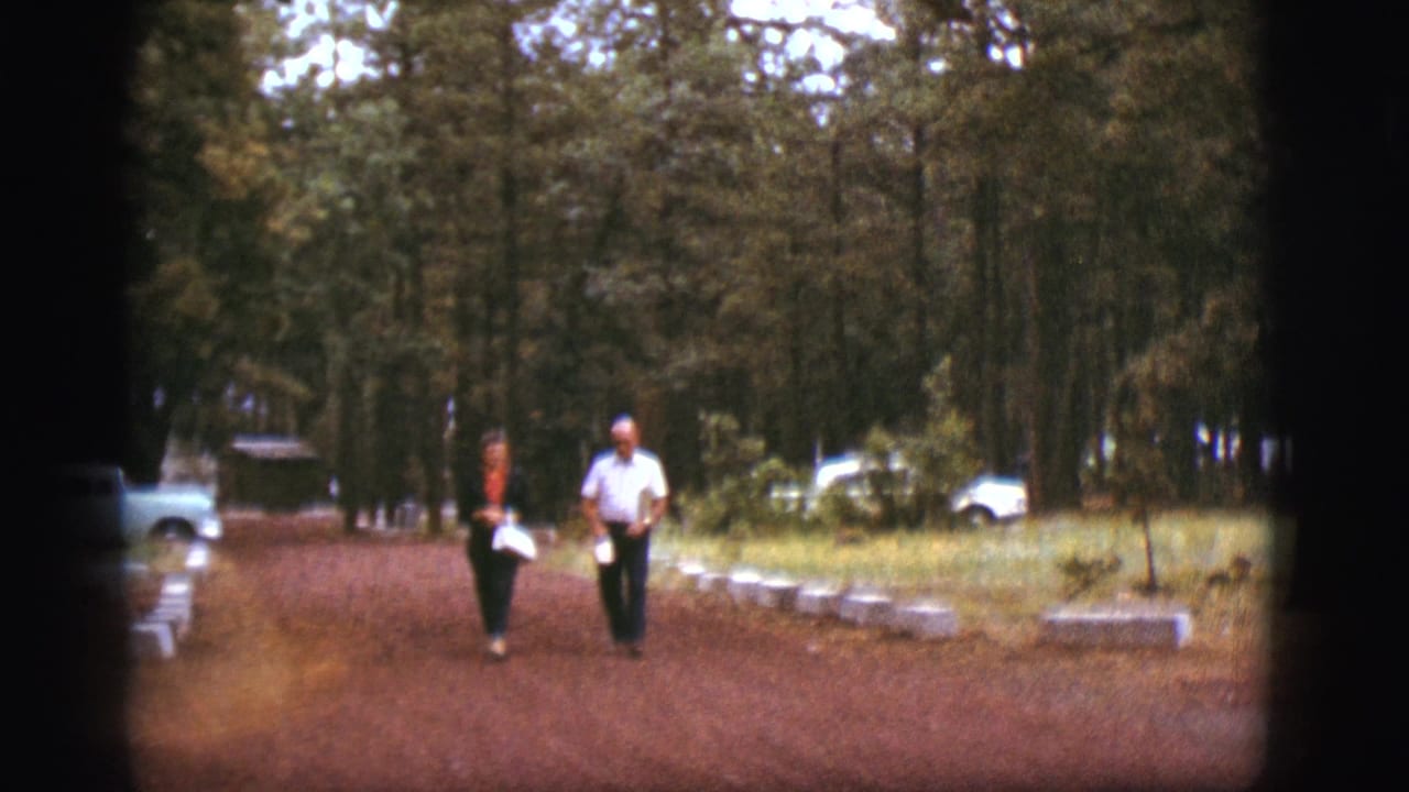 The couple walks closer together on the dirt path, with trees and parked cars behind them.