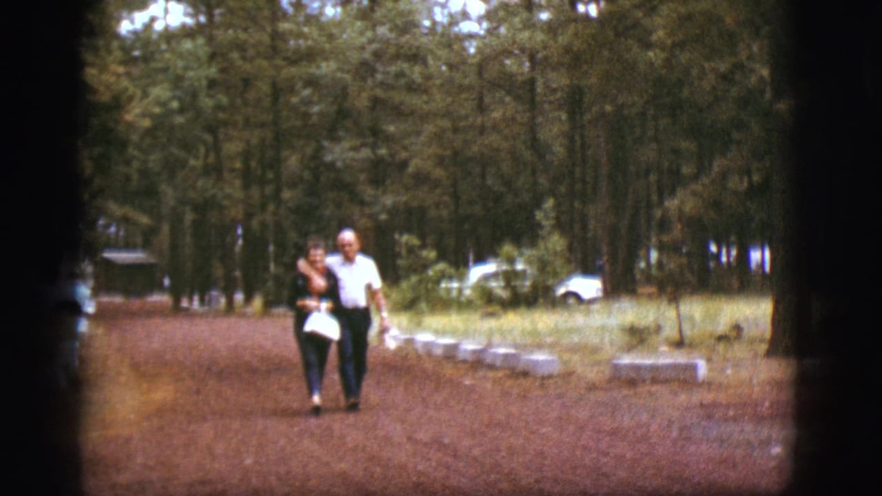 Elderly couple walking arm-in-arm on a dirt path through pine trees.