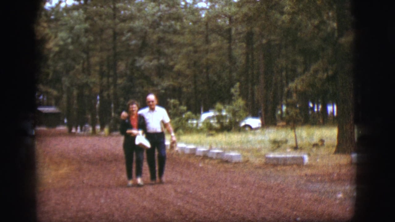 The couple continues walking arm-in-arm through the Flagstaff forest setting, with a white car behind the trees.