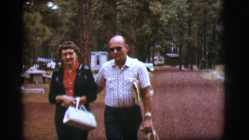 A smiling man in sunglasses and a woman carrying a white purse walk toward the camera on a wooded Flagstaff path.