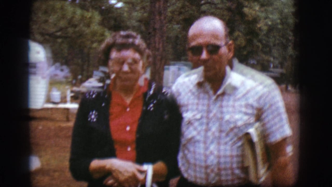 Two adults stand at a wooded campground near picnic tables.