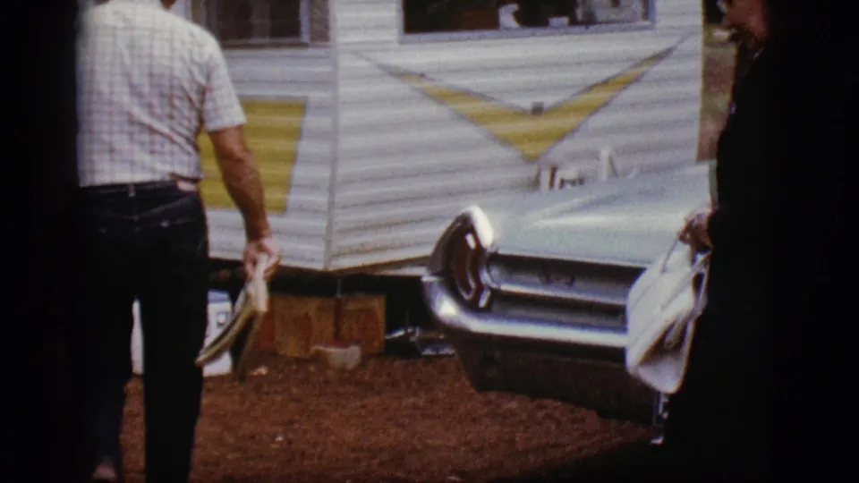 A man in a plaid shirt walks away near the white-and-yellow trailer and turquoise car in the 1968 Flagstaff campground footage.