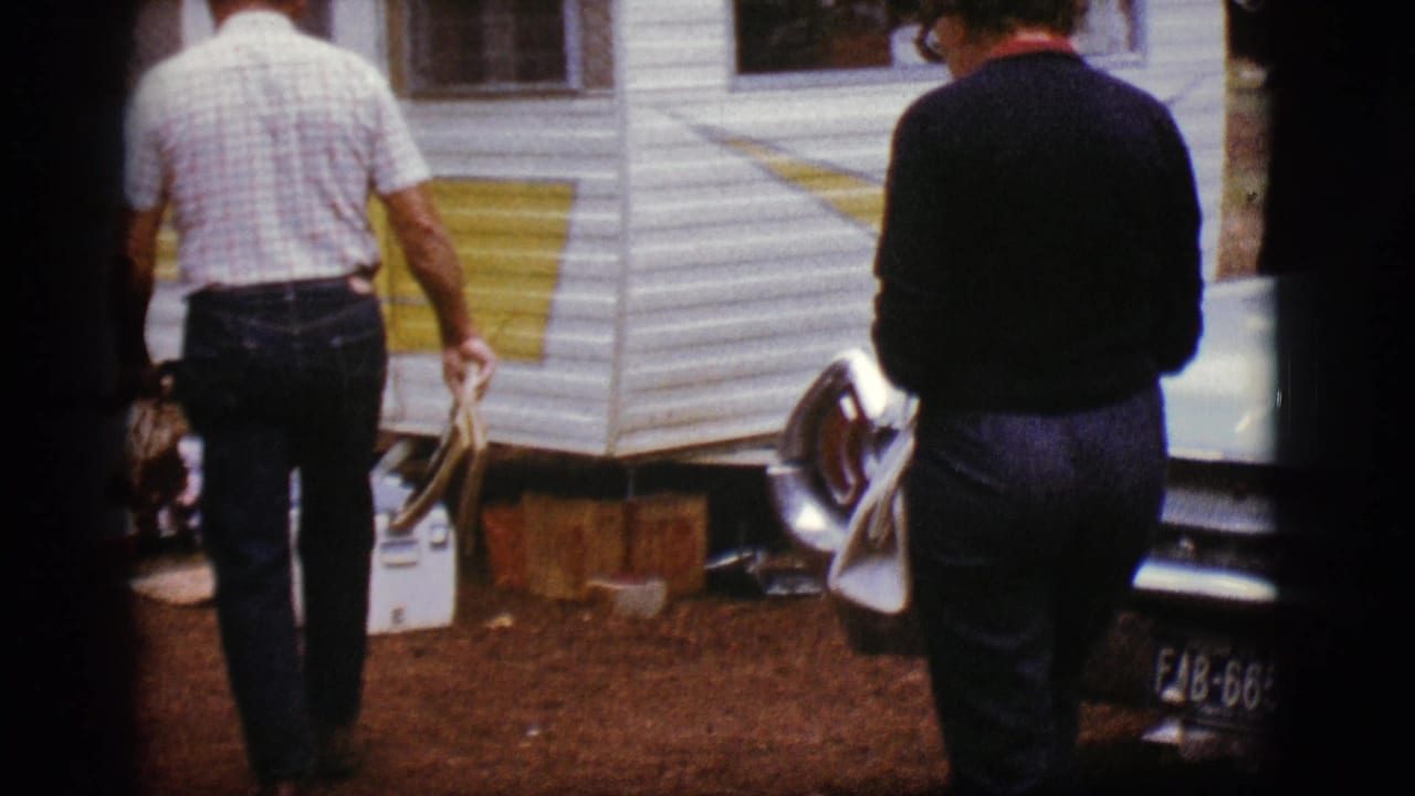 Older man walking toward a white and yellow camper trailer at a campsite.