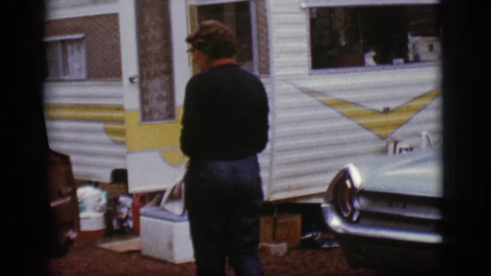 A man stands with his back to the camera beside the white and yellow camper trailer, with coolers and bags at the campsite.