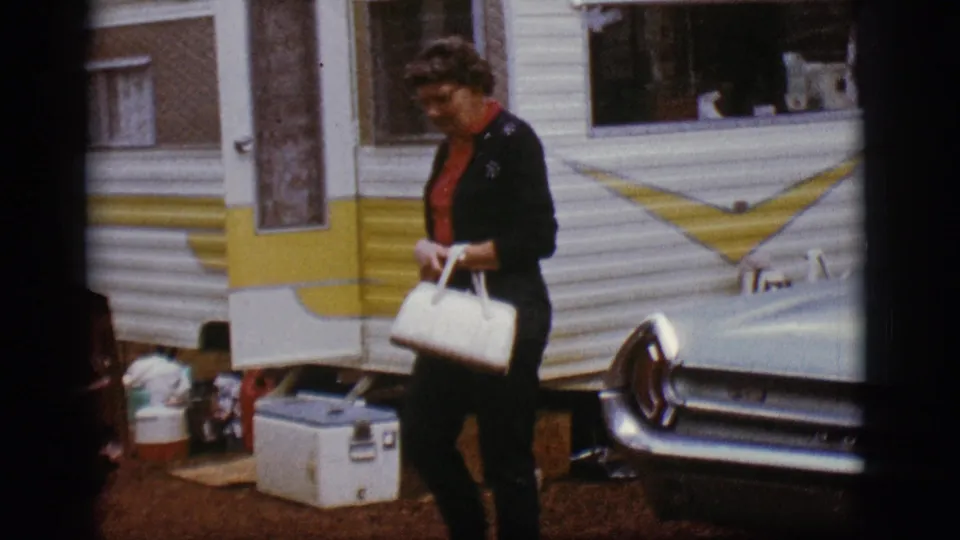 A woman with glasses and a white handbag walks beside the camper trailer and classic car in the 1968 Flagstaff campsite scene.