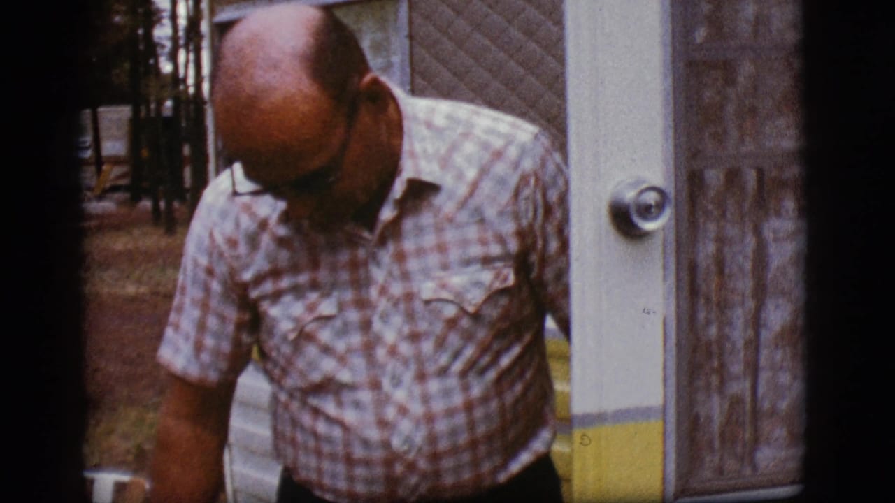A man in a plaid shirt looks downward at the doorway of a white and yellow camper trailer in the Flagstaff home-movie scene.