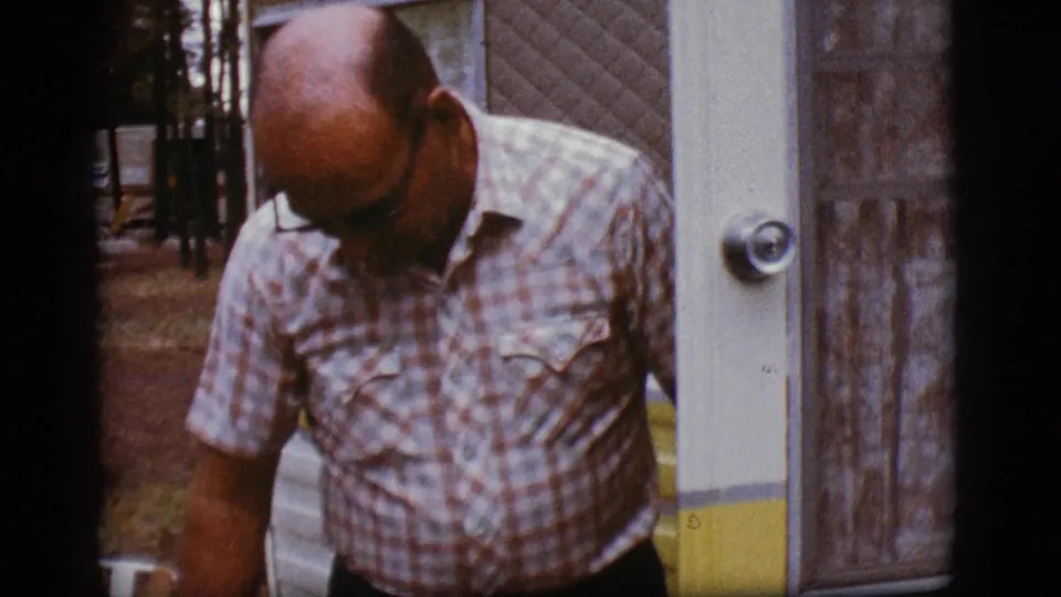 A man in a plaid shirt looks downward at the doorway of a white and yellow camper trailer in the Flagstaff home-movie scene.