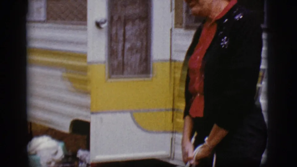 A woman in a dark sweater and red shirt passes beside the camper trailer while holding a white purse in the 1968 Flagstaff footage.