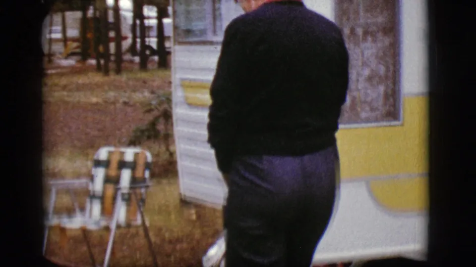 A person stands with their back to the camera near the camper trailer, with a folding chair and trees visible in the yard.