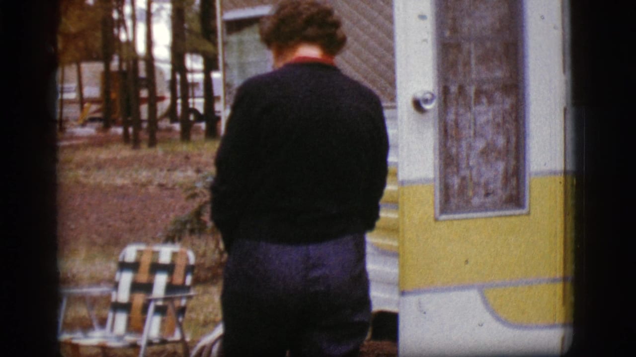 Woman seen from behind standing beside a white and yellow travel trailer in a wooded campground.