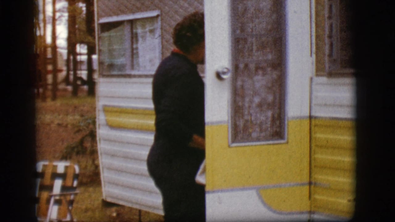 Woman in dark clothing stands partly turned at the travel trailer door near trees and a folding chair.