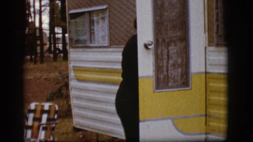 The woman moves partly behind the trailer doorway or side panel, leaving the yellow-striped trailer, chair, and wooded campsite prominent.