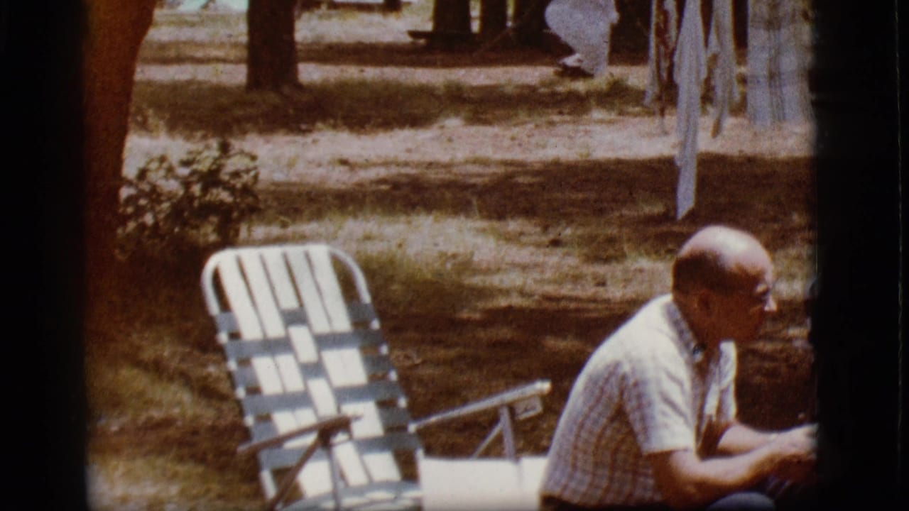 Man in glasses sits on a folding chair outside near trees and a clothesline.