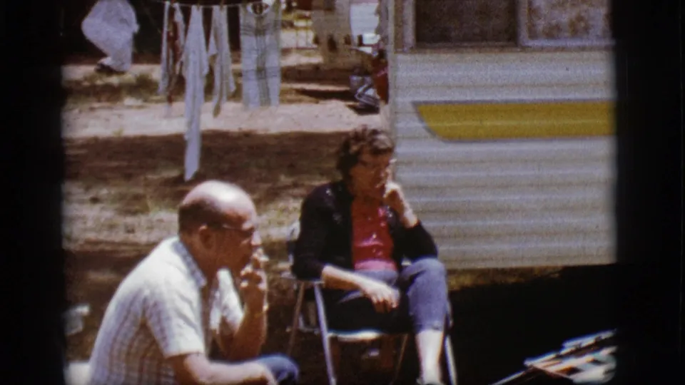 A woman in a dark cardigan and red top sits in a folding chair outside a trailer, with a man partly visible in the foreground.