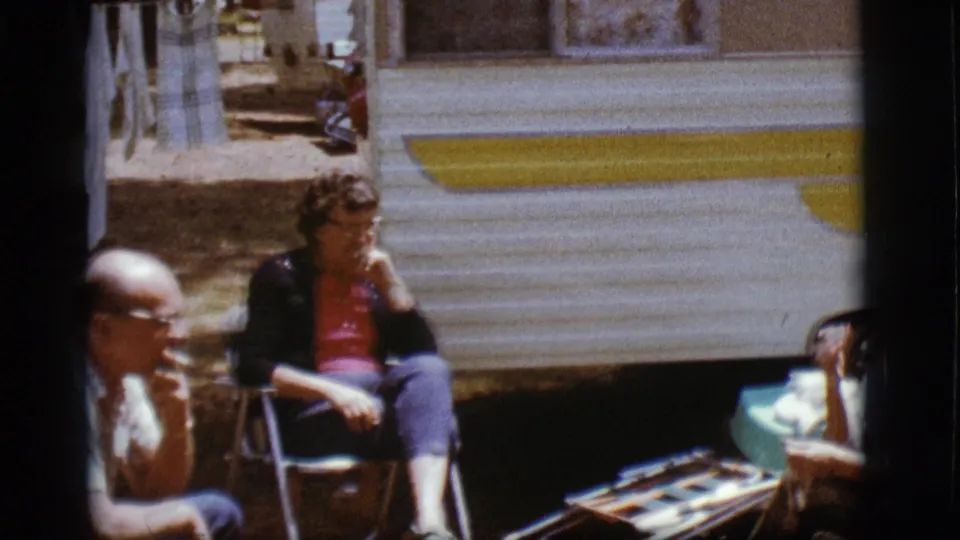 Two adults sit outside a white and yellow trailer in Flagstaff, with a third seated figure partly visible at the edge of the frame.