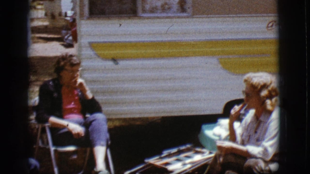 Two women seated outside a white and yellow travel trailer, eating and talking.