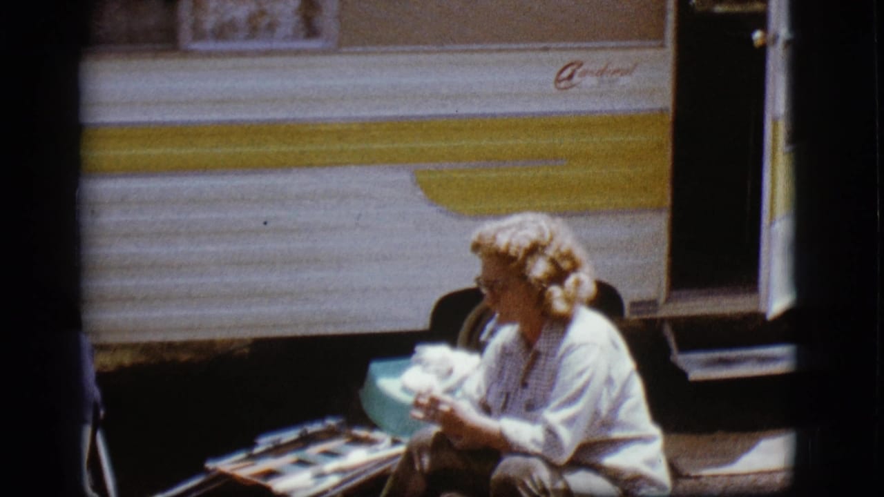 A woman sits near a light-blue basin beside the open door of a striped trailer.