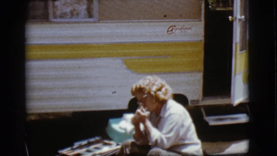 1968 Flagstaff frame of a woman seated outside a Cardinal RV, leaning over food in bright sunlight.