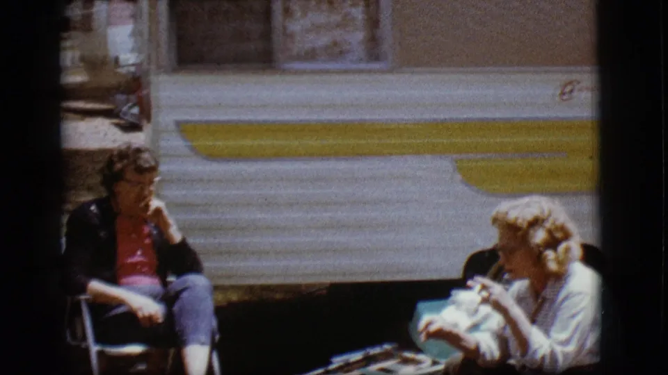 In 1968 Flagstaff, Arizona footage, two women rest in folding chairs outside a white trailer with yellow trim.
