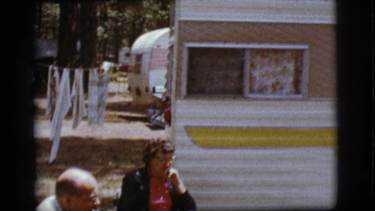 Two adults sit near a white and yellow camper with laundry hanging nearby.