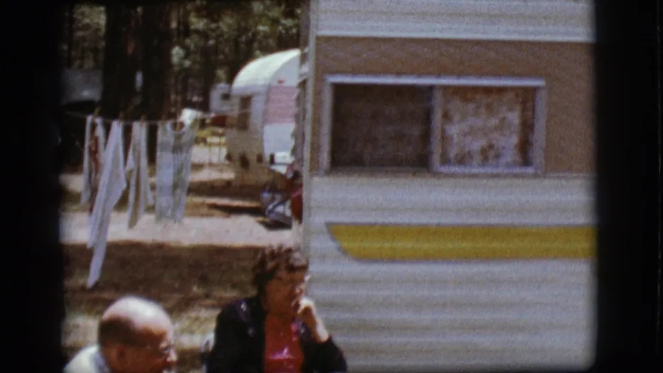 1968 Flagstaff home-movie frame showing seated adults beside a white and yellow camper at a wooded campground.