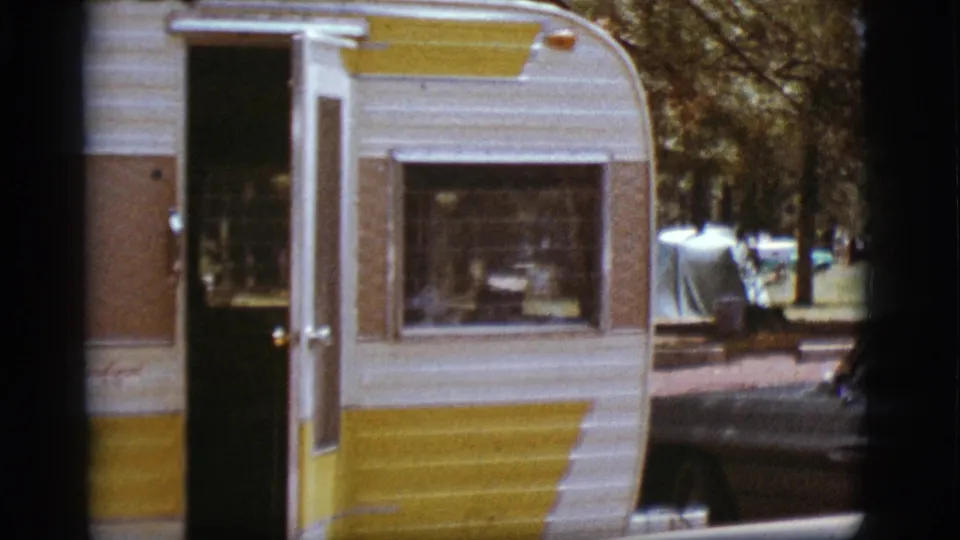 The camper fills most of the frame, with its open doorway, curtained window, tent, trees, and nearby car visible at the campground.