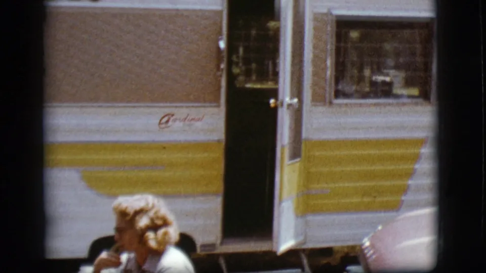1968 Flagstaff campground frame showing the striped camper and open door, with a blonde woman seated in the lower foreground.
