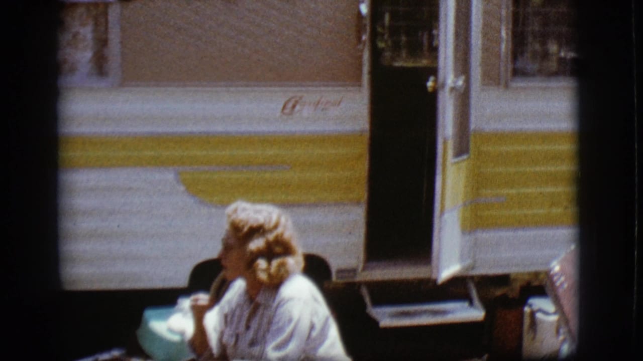 Blonde woman seated outside a white-and-yellow camper near an open door.