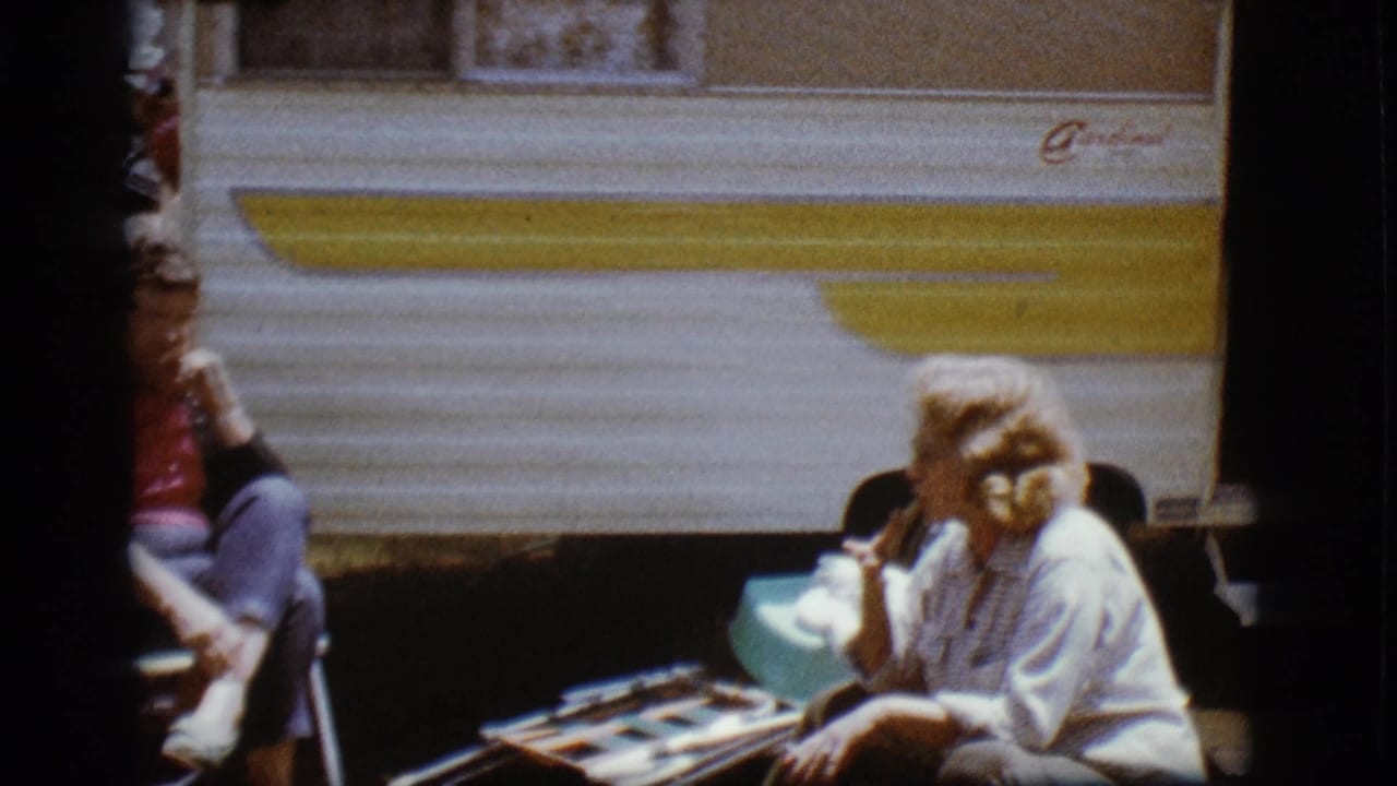 Woman seated beside a camper with another person partly visible nearby.