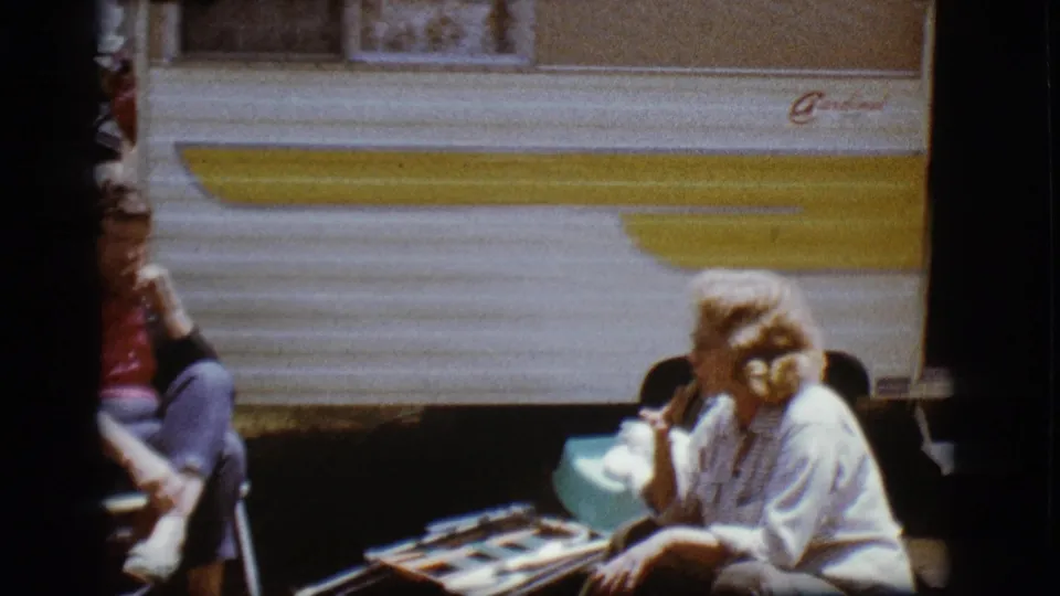 A woman sits beside the white-and-yellow Cardinal camper in Flagstaff, while another seated adult is partly visible at the edge of the frame.