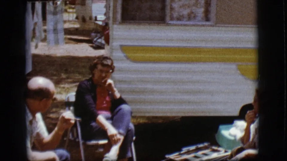 A man and a woman sit in folding chairs outside the camper in 1968 Flagstaff, with a second woman partly visible at the right edge.
