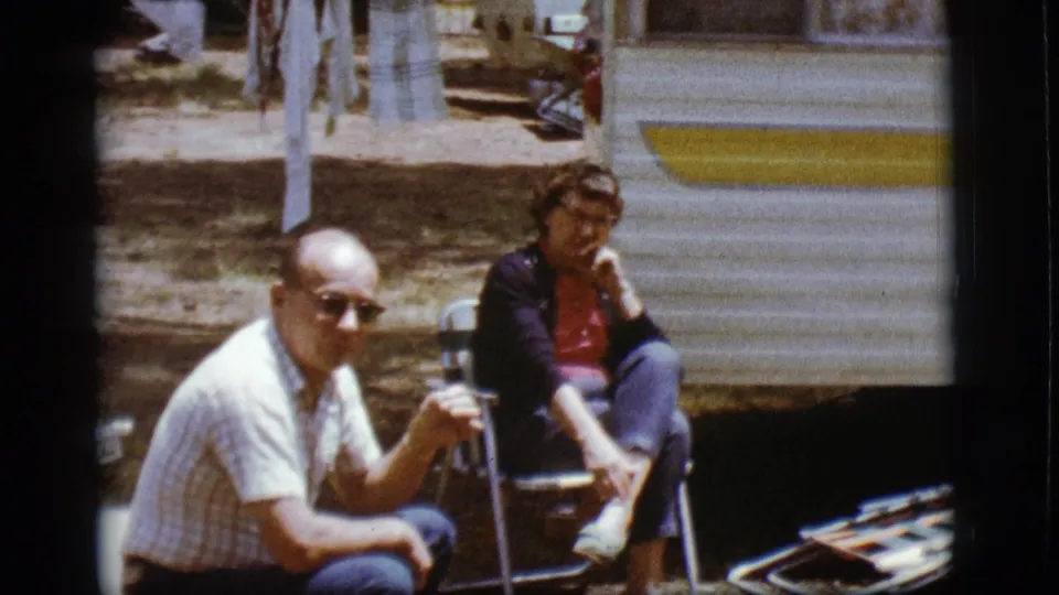 A man in sunglasses and a woman sit outside the white-and-yellow camper in Flagstaff, with folding chairs and travel items nearby.