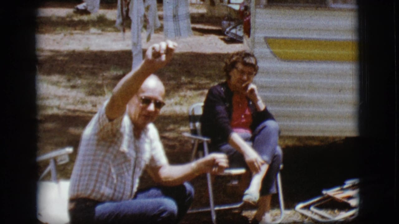 Older man waving beside seated woman outside a travel trailer at a campsite.
