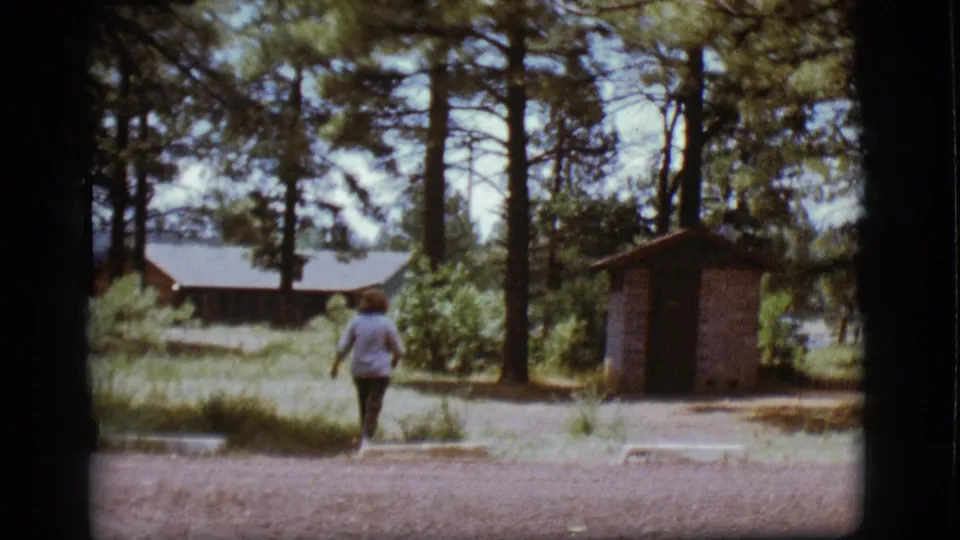 The walker continues away through the Flagstaff campground, framed by tall pines, a low building, grass, and a small utility structure.