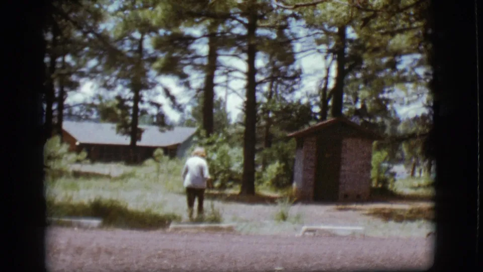 A light-shirted adult walks away in the 1968 Flagstaff campground scene, with pine trees and modest camp structures in view.