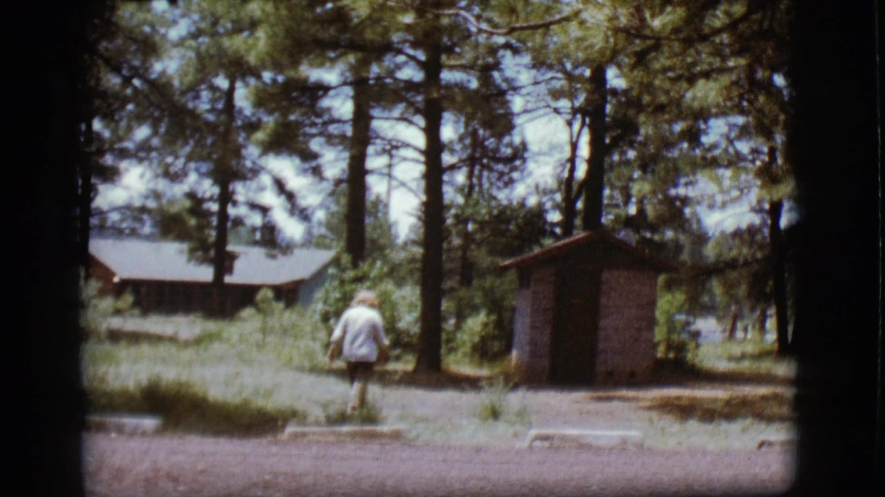 1968 Flagstaff home-movie frame showing a woman walking across a gravel campsite with pine trees, a cabin, and an outhouse nearby.