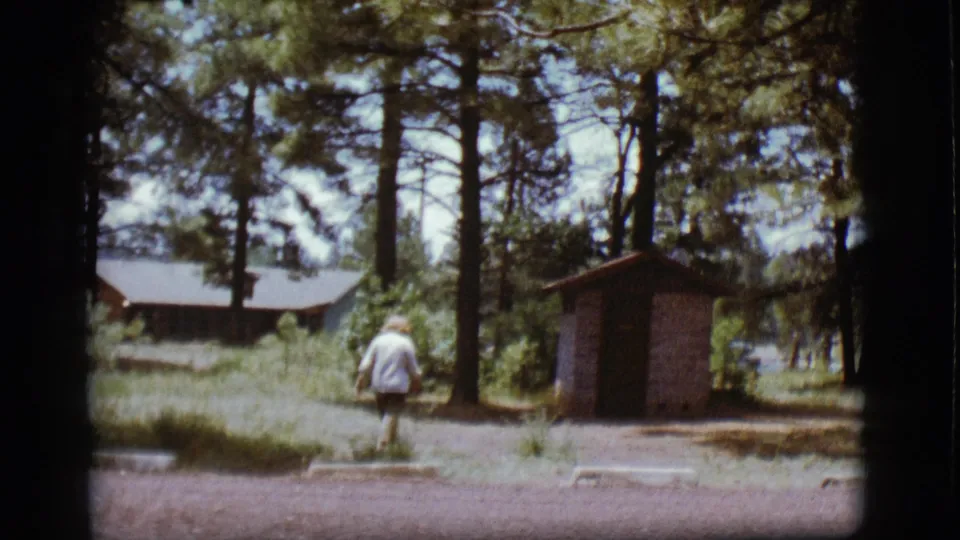 1968 Flagstaff home-movie frame showing a woman walking across a gravel campsite with pine trees, a cabin, and an outhouse nearby.
