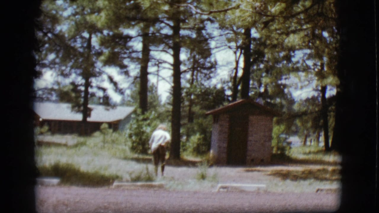 Woman farther along a gravel path beside pine trees and a small outhouse.