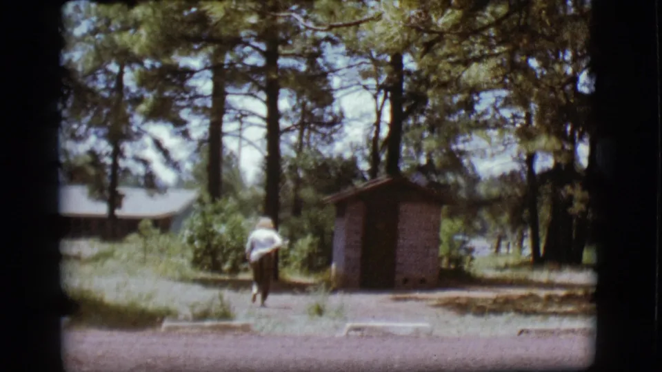 1968 Flagstaff home-movie still showing the woman approaching the small outhouse among tall pine trees and open campground groundcover.