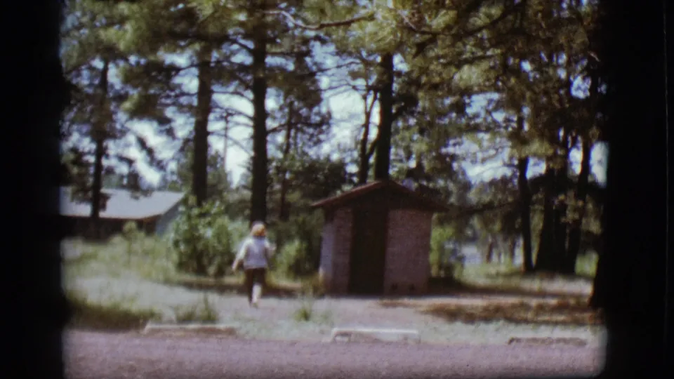 1968 Flagstaff frame showing the woman close to the small brick outhouse, with pine trees, a cabin, and gravel campground space around her.