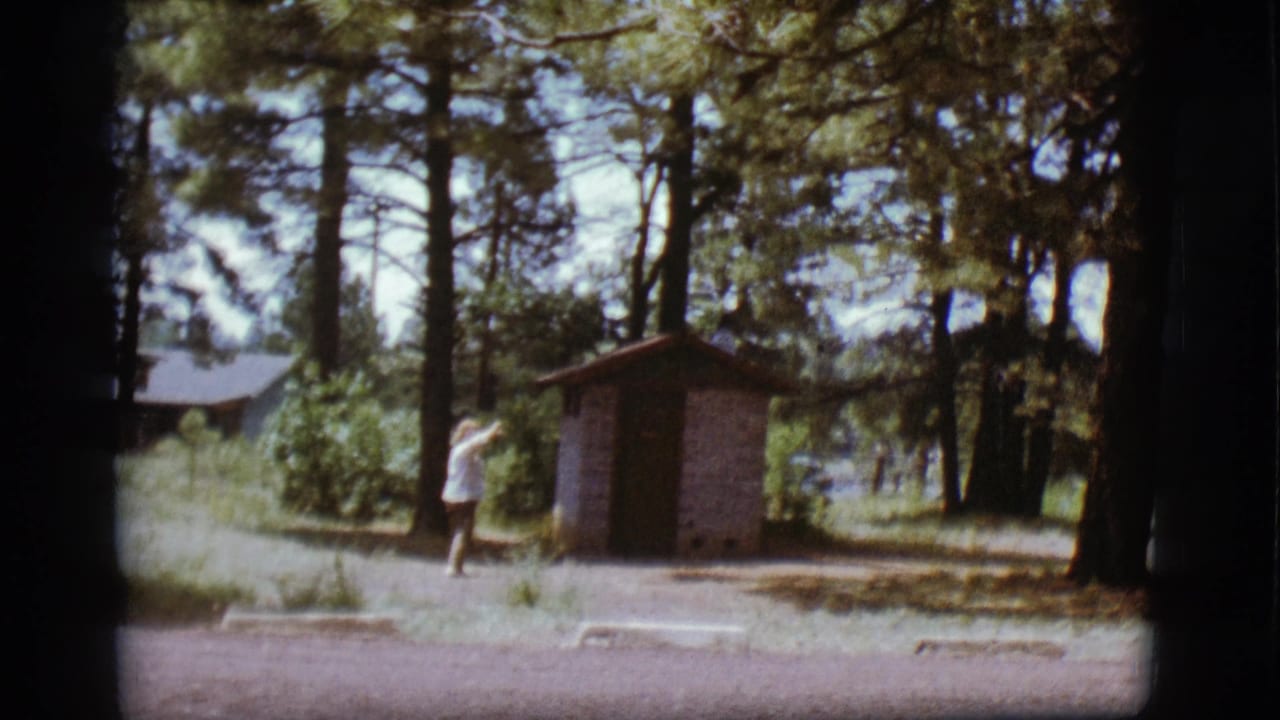 1968 Flagstaff home-movie frame showing the woman near shrubs and a brick outhouse under tall pines.
