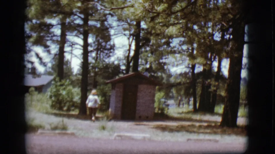 1968 Flagstaff 8mm still with the woman walking away from camera toward a small brick outhouse in a shaded pine setting.