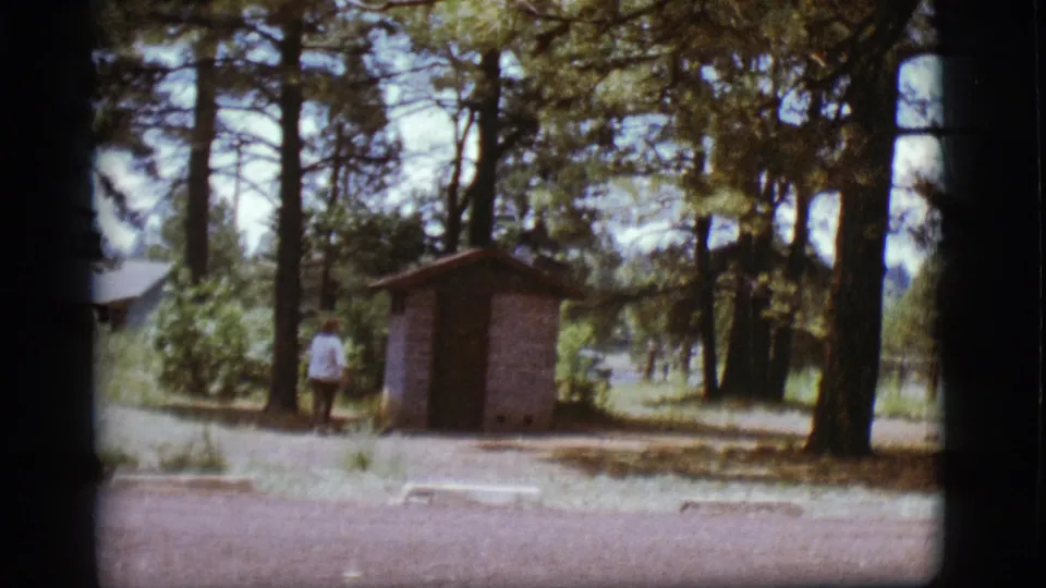 1968 Flagstaff frame of a woman near a brick outhouse, surrounded by tall pines, low plants, and bright midday light.