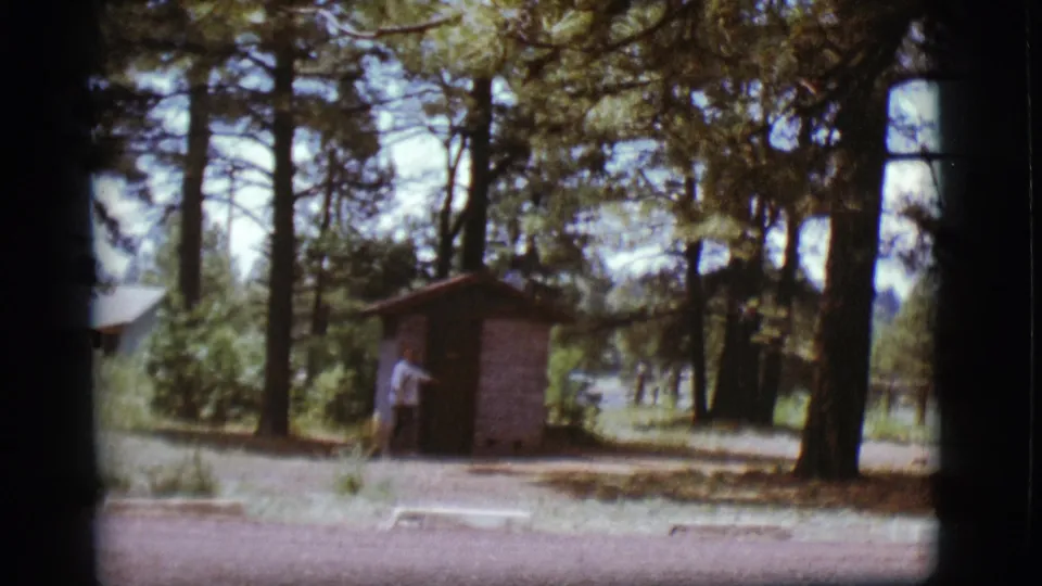 1968 Flagstaff-area frame with a woman gesturing toward a small shed door beneath pine trees.